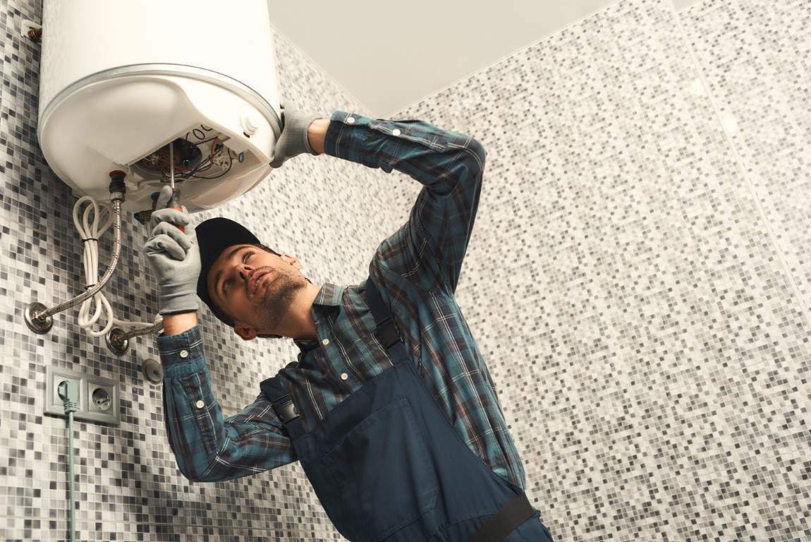 Plumber installing electric heating boiler. Young handyman in uniform setting up electric heating boiler at home